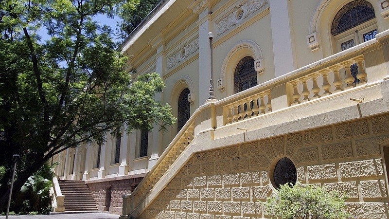 Fachada do prédio do Arquivo Público do Rio Grande do Sul. com arquitetura do começo do século XX, paredes amarelas, pilastras brancas e grandes janelas em arco. O acesso a entrada se dá por duas escadarias externas, uma para direita e outra para esquerda, construídas em cima de uma base de pedras, a mesma utilizada no prédio. A área externa é arborizada e agradável.