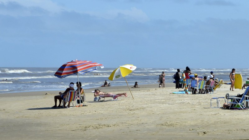 Imagem da beira da praia, em um dia de céu azul, com pessoas sentadas em cadeiras, algumas debaixo de guarda-sol e algumas crianças brincando na areia.