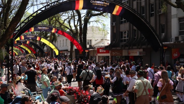 Foto de pessoas vestidas com roupas típicas alemã, desfilando por uma rua na Oktoberfet. Há arcos sobre a rua, enfeitados de preto, amarelo e vermelho - cores do evento. Várias pessoas sentadas na calçada assistindo a passeata.