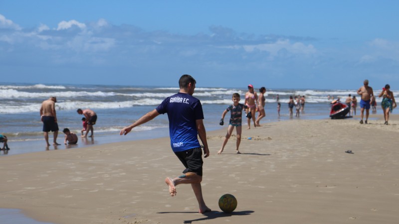 Foto da beira da praia. No centro da imagem um homem de camisa azul se prepara para chutar uma bola de futebol. Ao redor banhistas aproveitam o dia ensolarado.