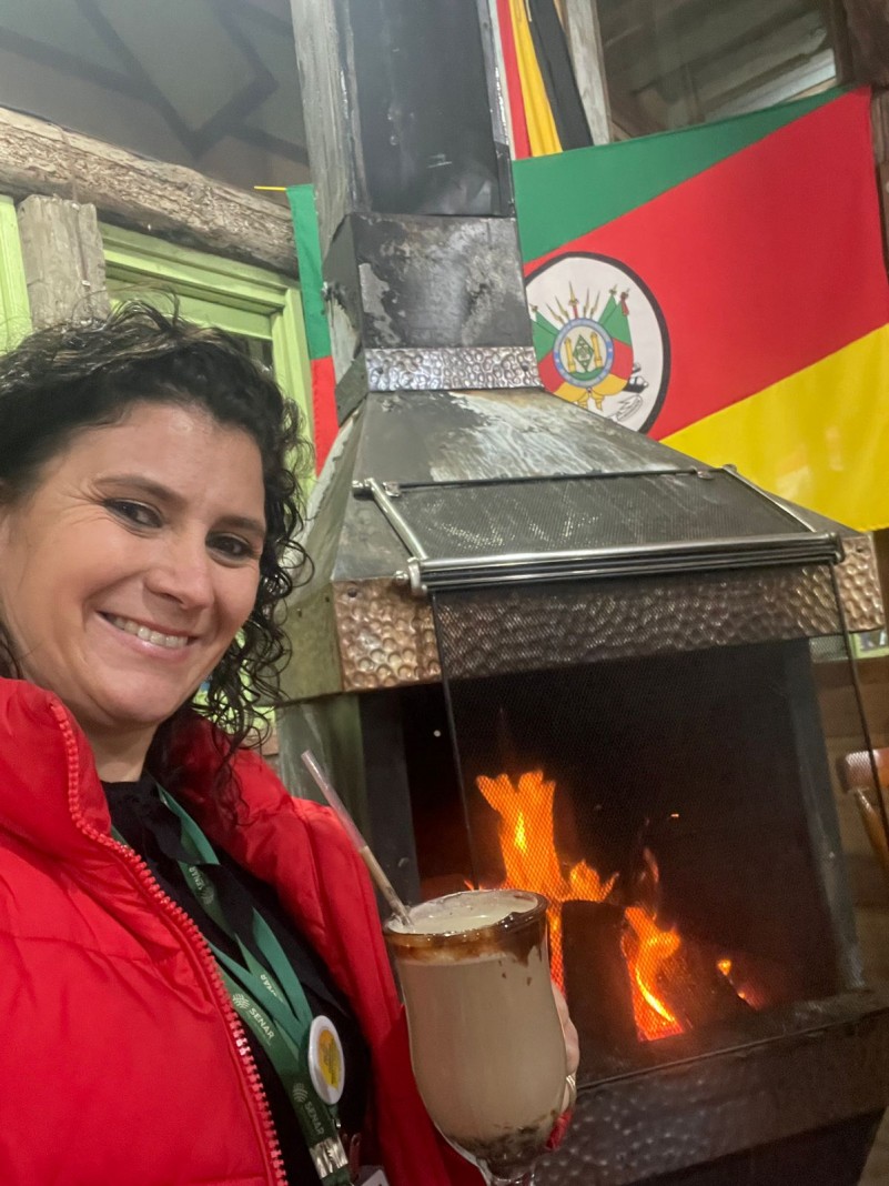 claudiana aparece com roupa de frio e uma bebida quente em frente a uma churrasqueira