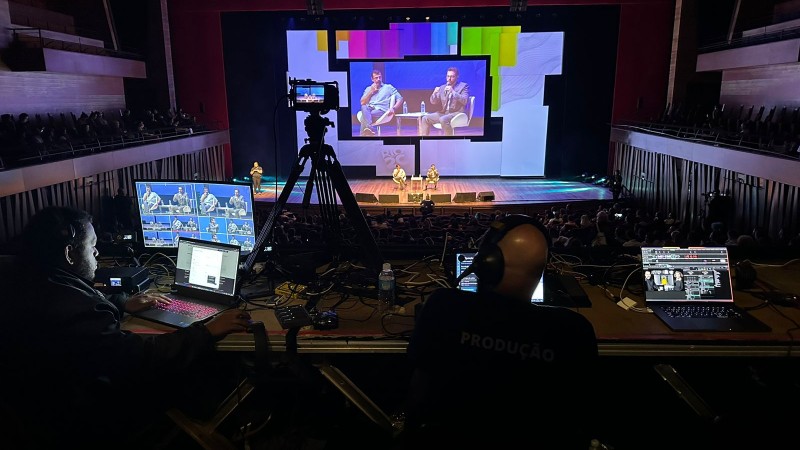 Operadores trabalham durante painel na Rio 2C. Na foto é possível ver um palco com um telão colorido e vários monitores com seus computadores. Há câmeras focando nos dois palestrantes presentes no palco.