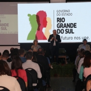 Plateia contemplando a fala do Secretário de Turismo. Telão exibe o logo do estado do Rio Grande do Sul ao fundo.