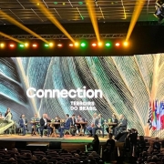Fotografia colorida do palco de um audit&oacute;rio durante um painel do evento "Connection Terroirs do Brasil". No fundo do palco, um enorme painel de LED exibe uma textura de folhas verdes em tons claros e escuros, com o nome do evento escrito em letras brancas no centro. Diante do tel&atilde;o, cerca de dez participantes do painel, em trajes formais, est&atilde;o sentados em cadeiras de madeira dispostas em semic&iacute;rculo. 