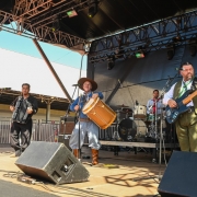 Fotografia colorida, em plano m&eacute;dio e &acirc;ngulo de baixo para cima, mostra um grupo musical masculino de m&uacute;sica tradicionalista ga&uacute;cha se apresentando em um palco sob a luz do dia.No palco, cinco m&uacute;sicos vestem trajes t&iacute;picos.